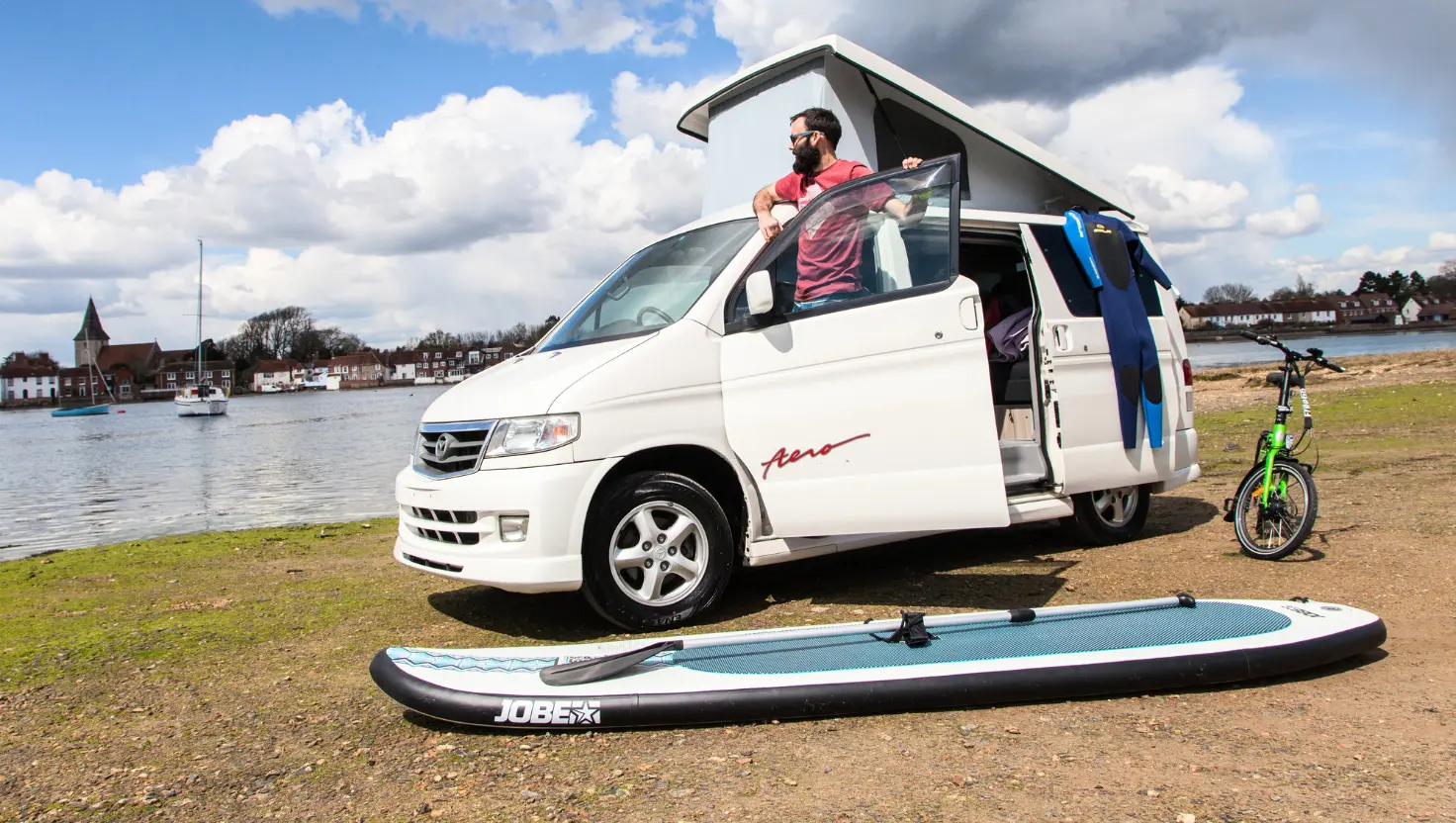 man standing at the door of a white campervan with a paddleboard next to the van for surfing campervan trips UK