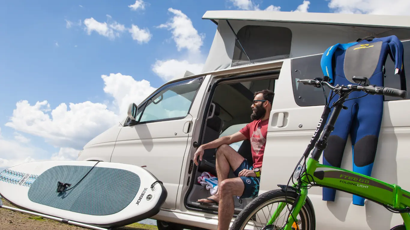 man sat in a white campervan with a bike and paddle board